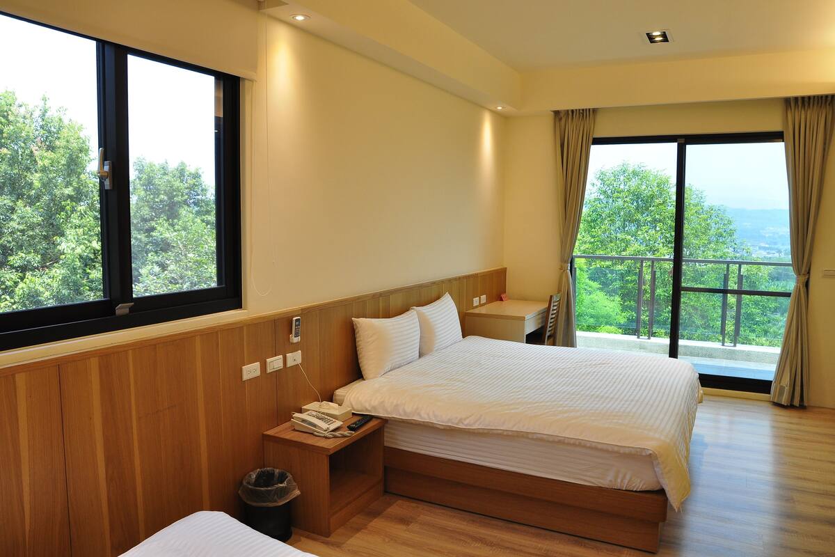 A hotel room with a large bed, wooden paneling, a small bedside table with a telephone, and a view of greenery through large windows.
