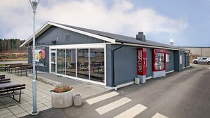 A modern building with a large glass entrance, outdoor seating, and a red sign advertising a restaurant. - Drive-in Motell (Mjolby)