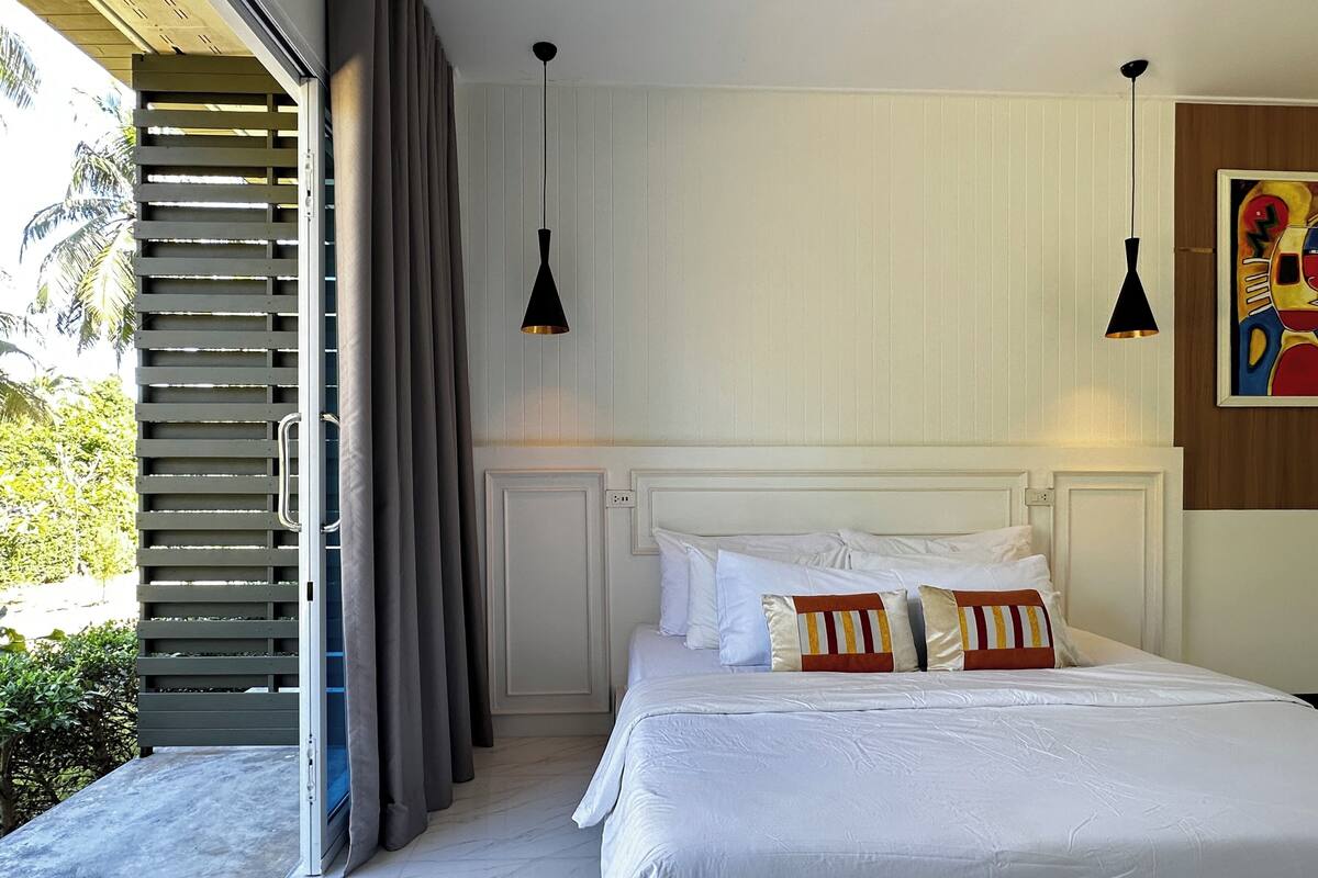 Un dormitorio con una cama grande, ropa de cama blanca y vistas a palmeras a través de un ventanal corredera.