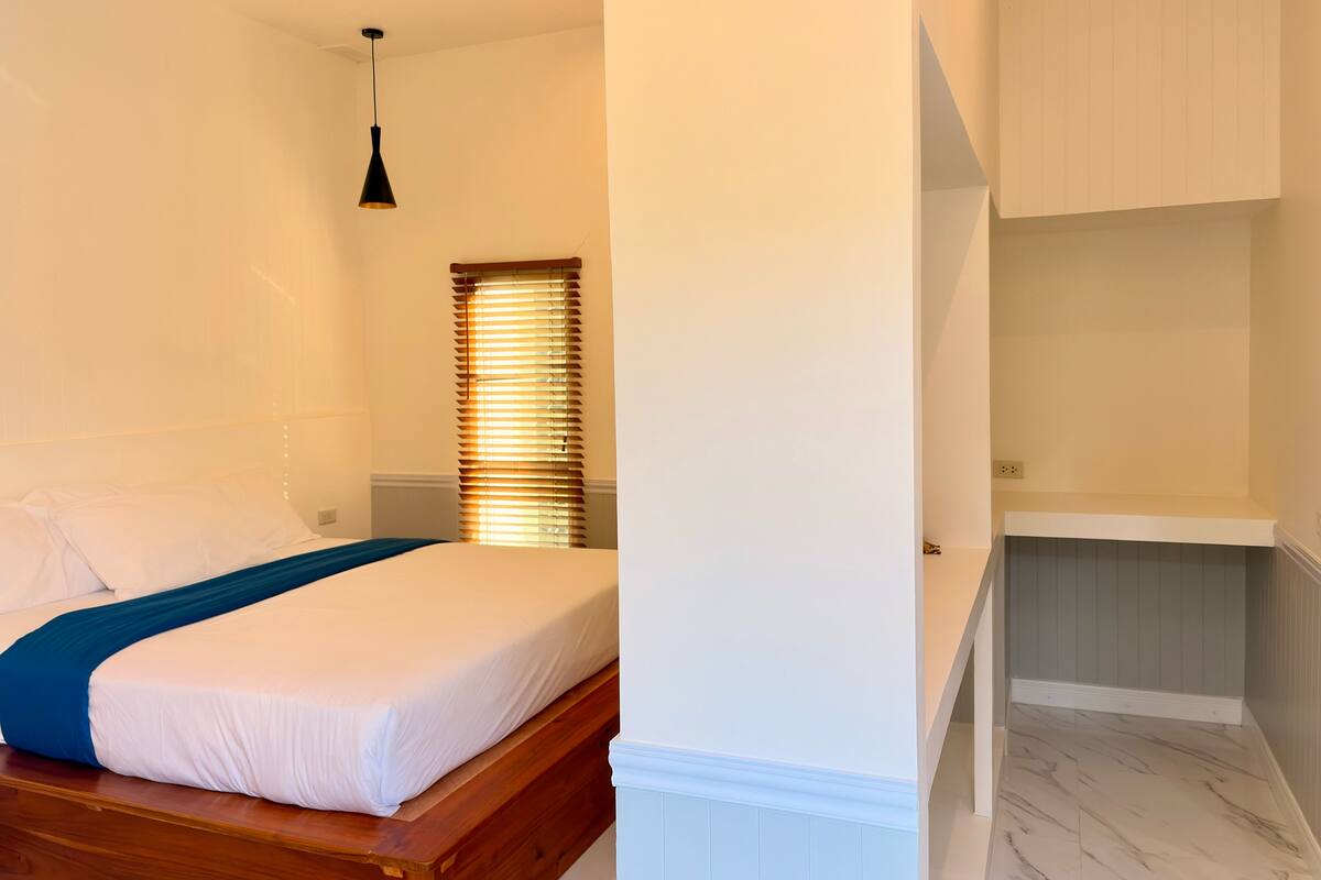 Un dormitorio con una cama de madera, paredes blancas, una ventana con persianas y una pequeña estantería.