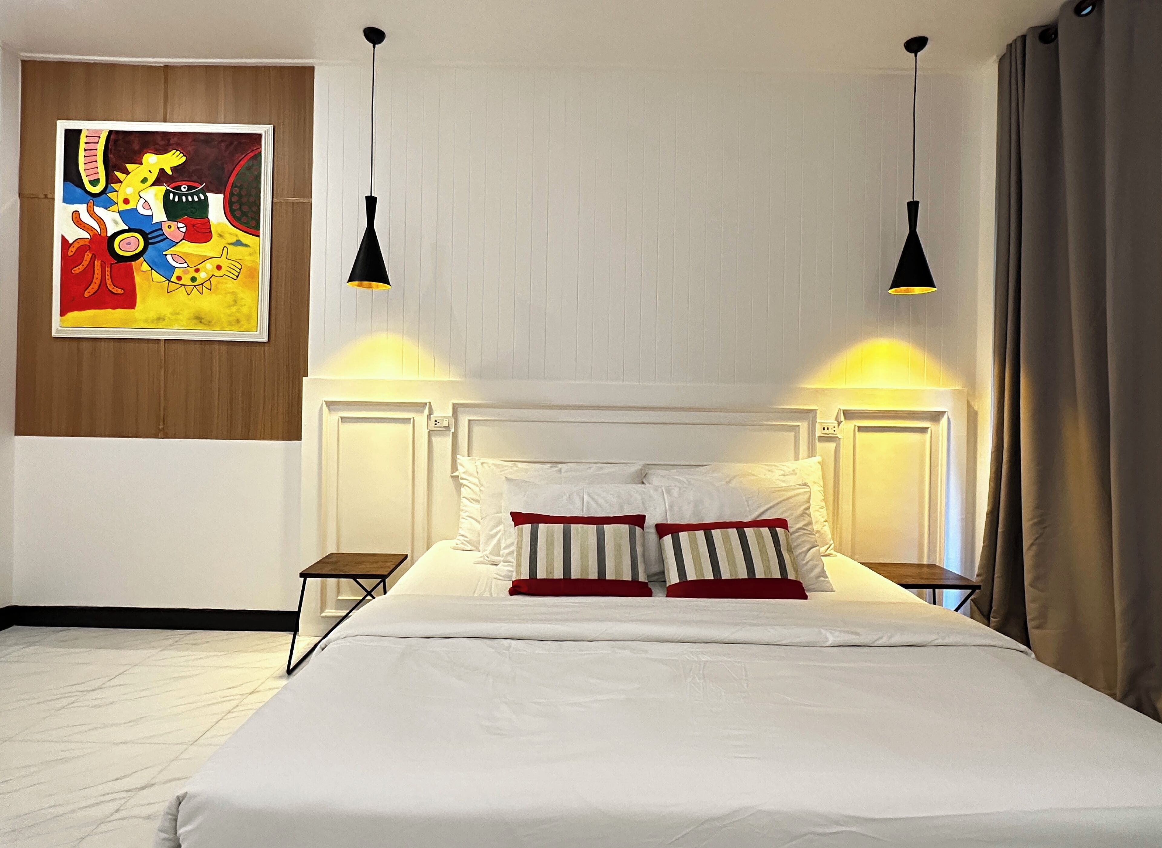 A neatly made bed with white linens and striped pillows, two bedside tables with lamps, a wooden panel wall, and a colorful abstract painting above the bed.