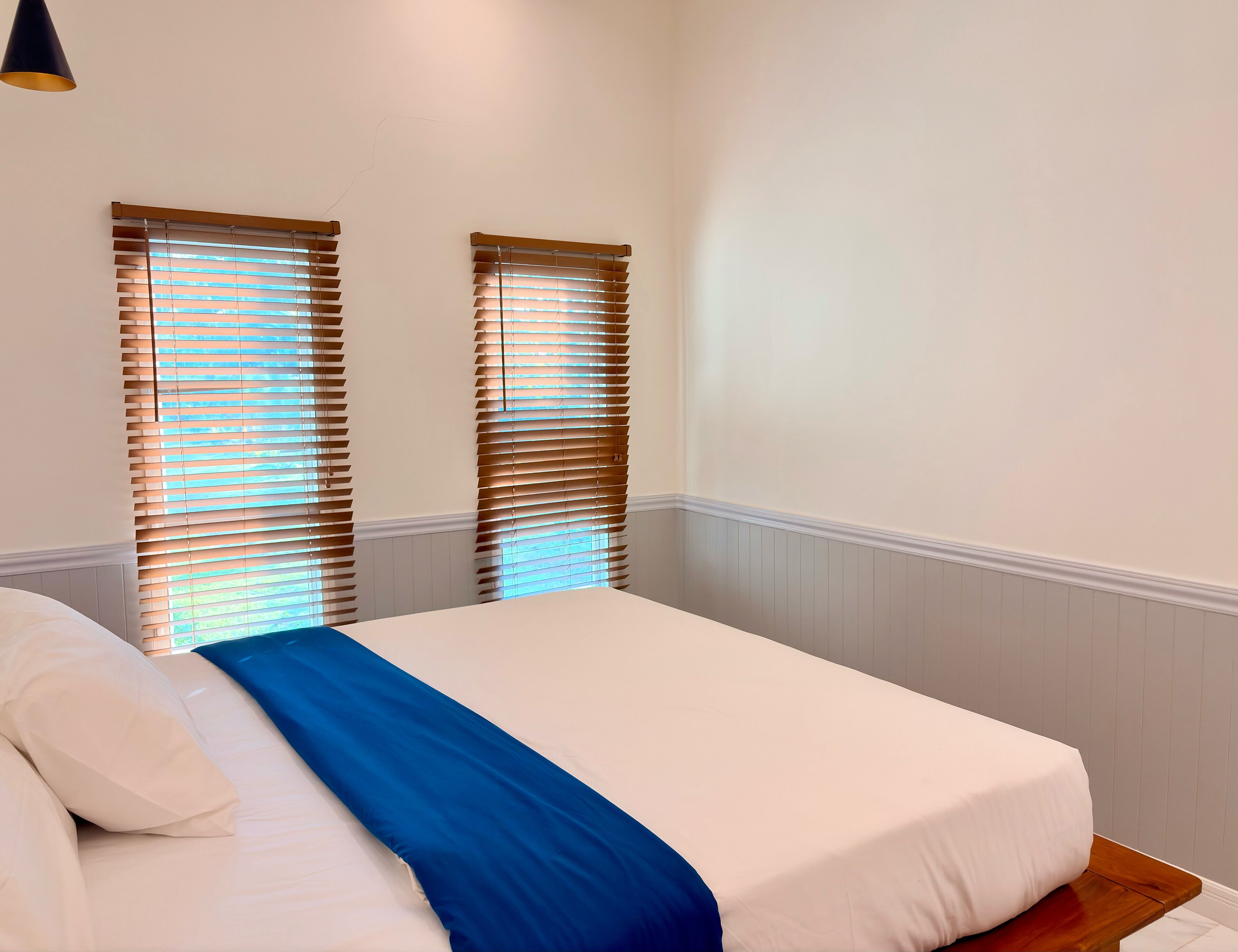 Ein Schlafzimmer mit einem Bett, weißen Wänden, Holzvertäfelung und zwei Fenstern mit Jalousien.
