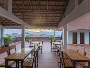 A covered outdoor seating area with wooden tables and chairs, potted plants, and a view of buildings in the distance. - Emily and Nana Lodge (Siem Reap)