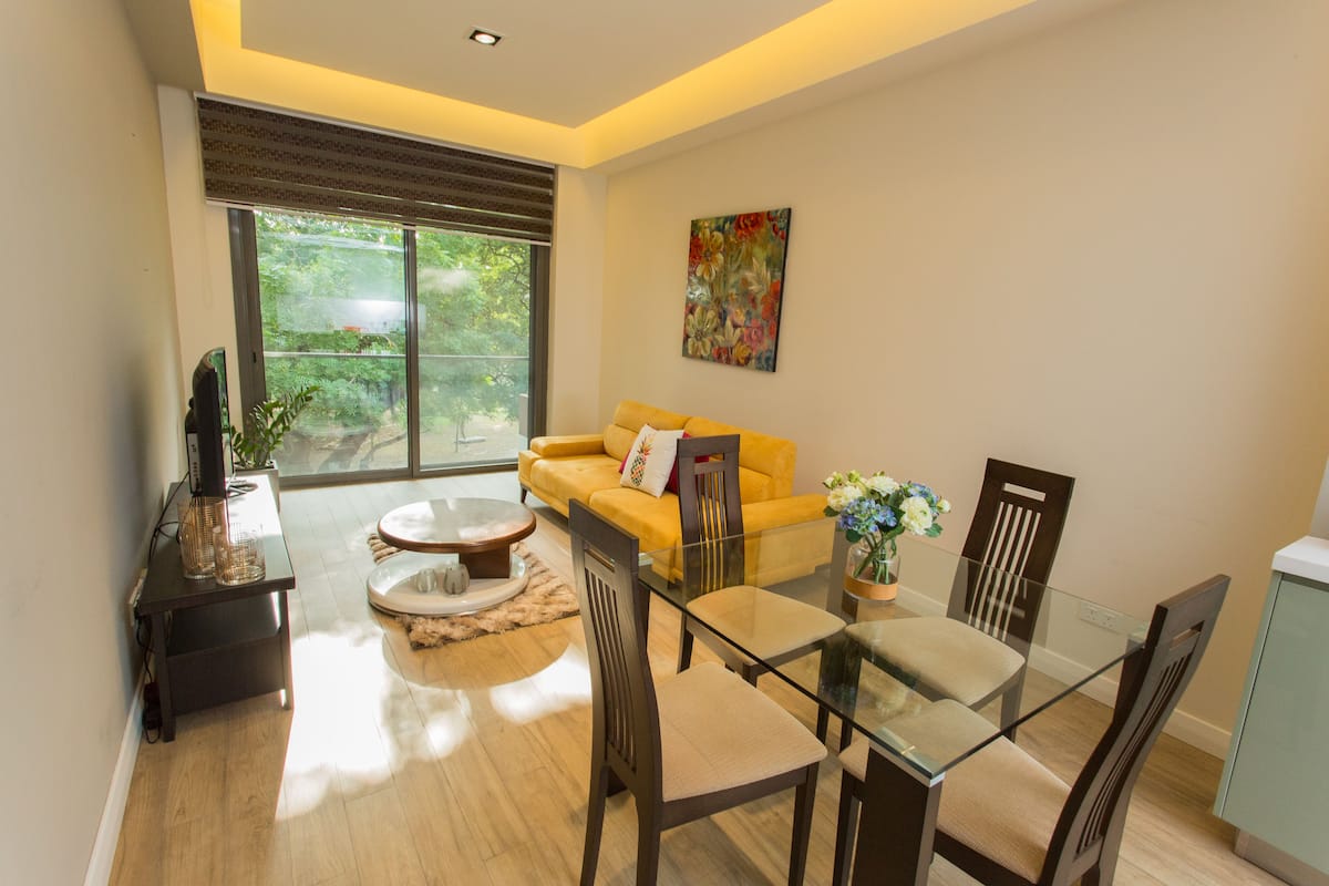 Een moderne woonkamer met een gele bank, een glazen eettafel en houten stoelen. Er is een groot raam met uitzicht op groen.