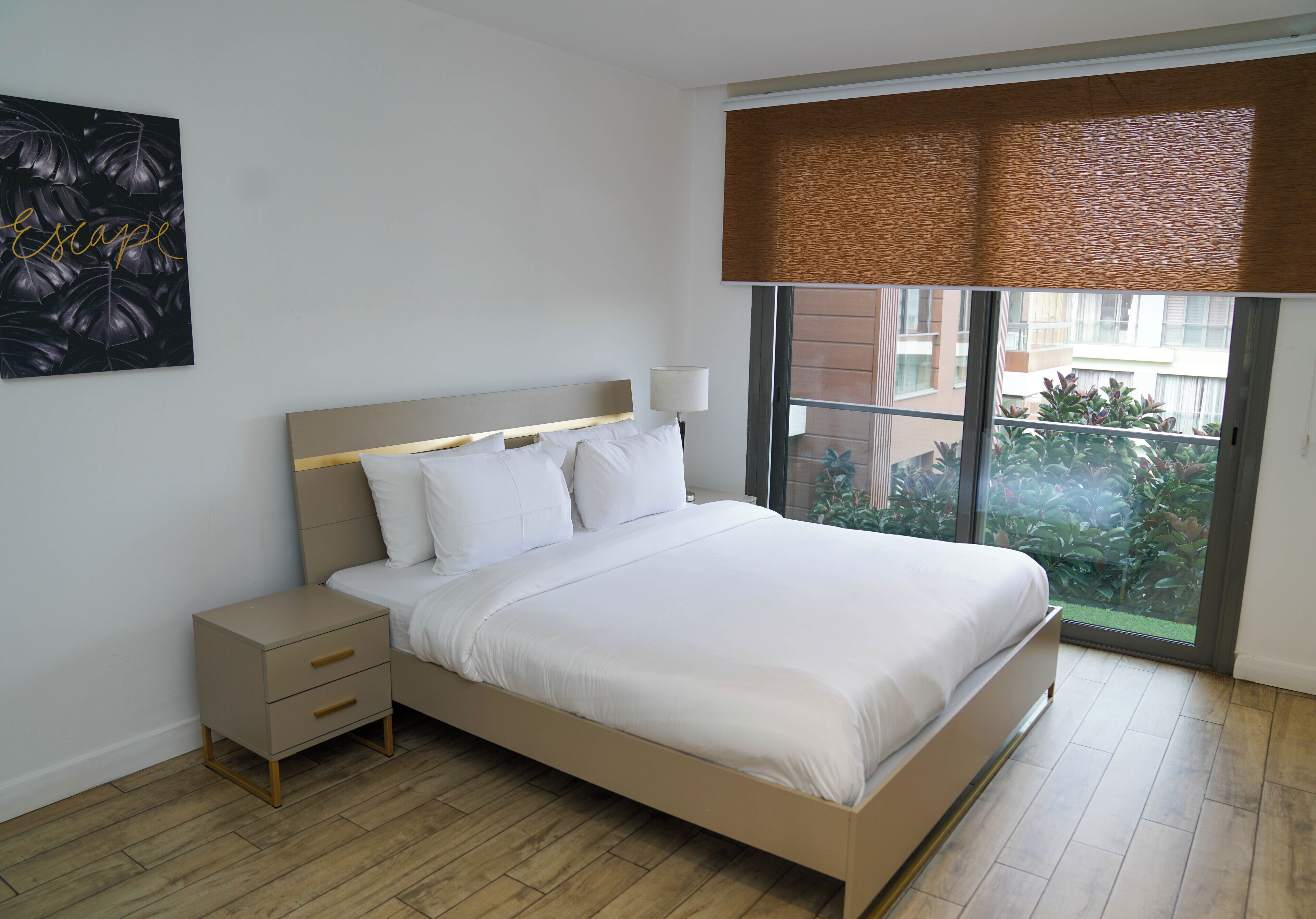 A modern bedroom with a large bed, a nightstand, a wall-mounted lamp, and a window with a view of buildings and greenery.