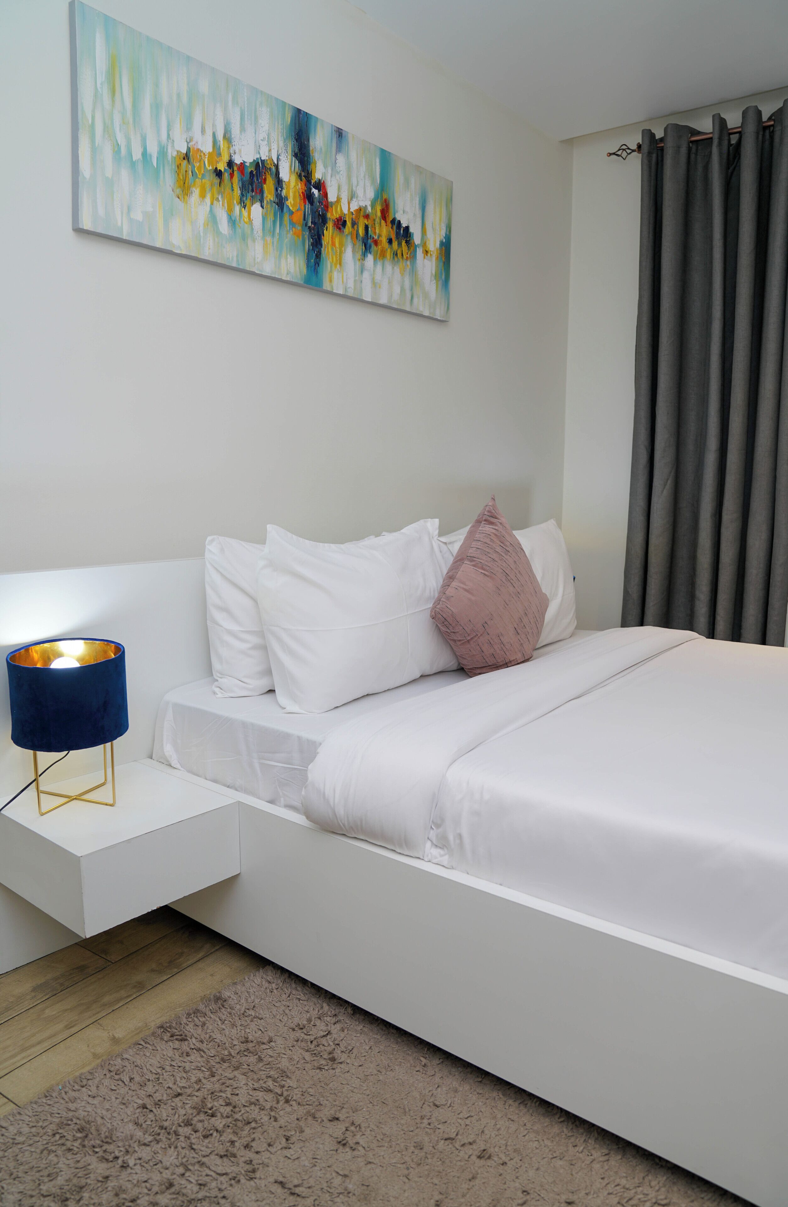 Ein modernes Schlafzimmer mit einem weißen Bett, einer blauen Nachttischlampe, einem flauschigen Teppich und einem Bild an der Wand.