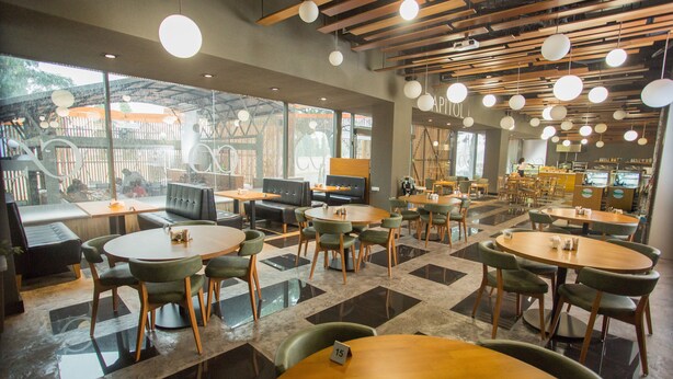 Um café moderno com mesas redondas e cadeiras verdes, janelas amplas e teto de madeira.