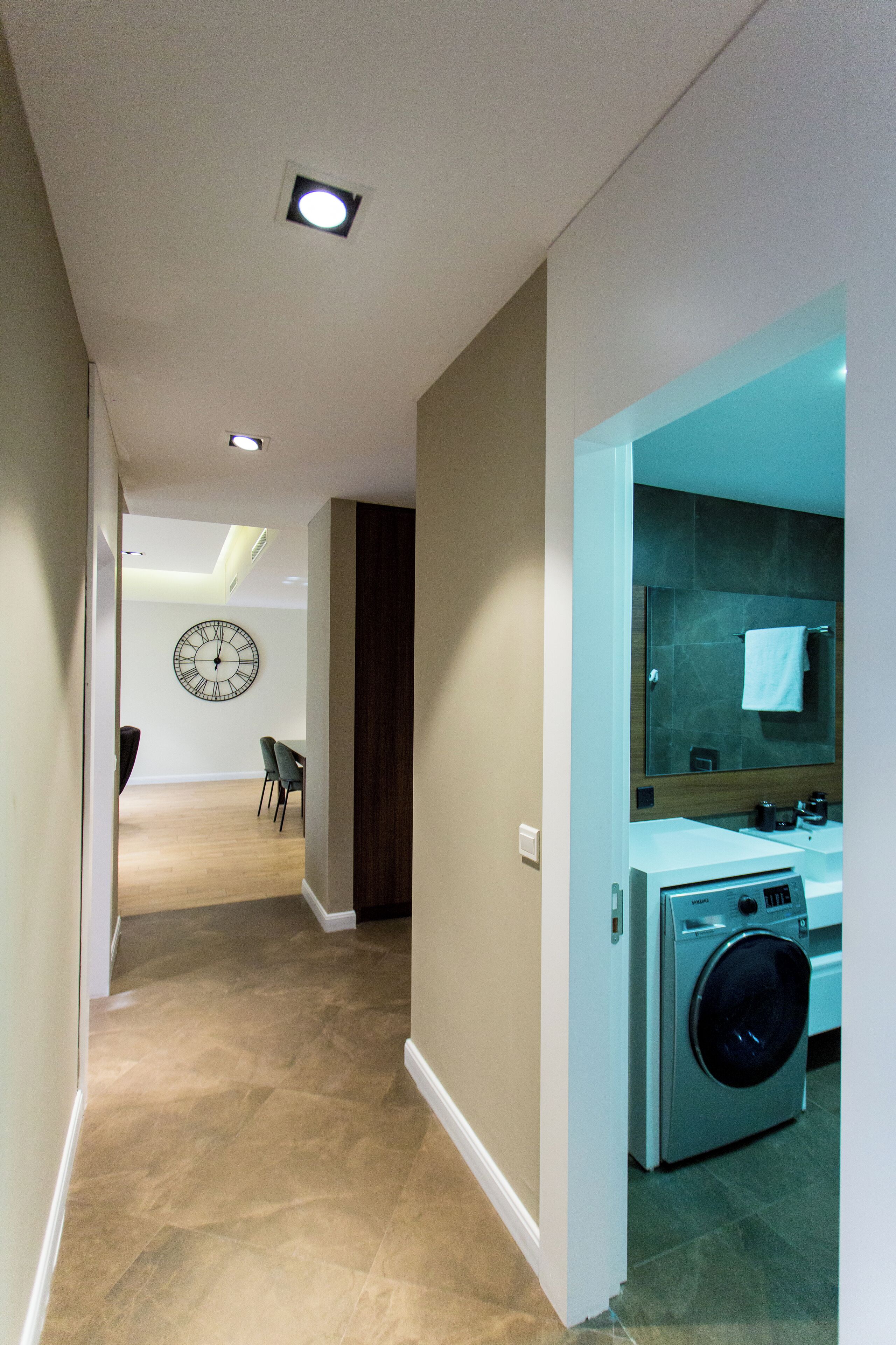 A modern kitchen with a built-in washing machine, a large wall clock, and a dining area with chairs.