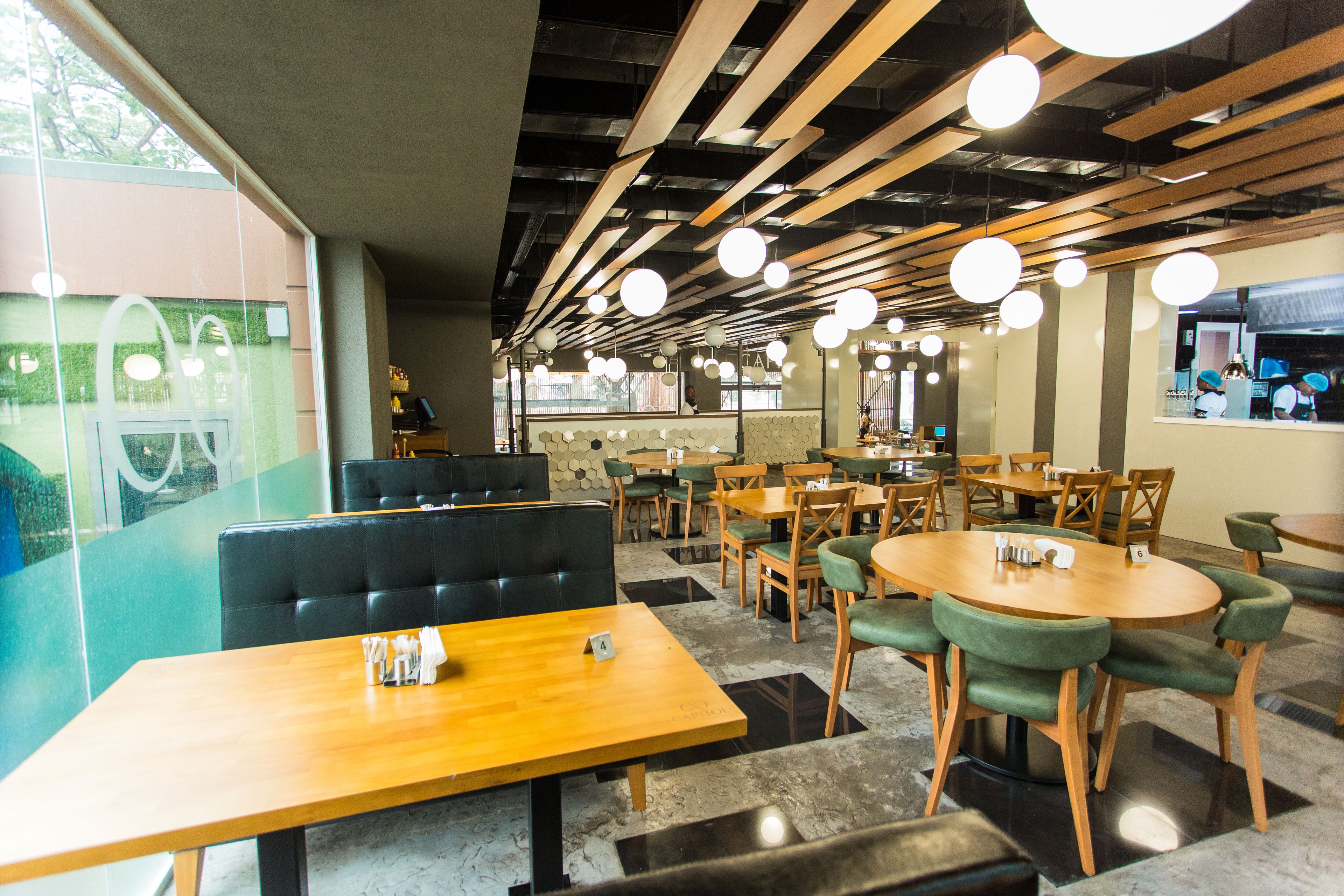 Ein modernes Restaurantambiente mit Holztischen, grünen Sitzgelegenheiten und einer hohen Decke mit freiliegenden Balken und Pendelleuchten.