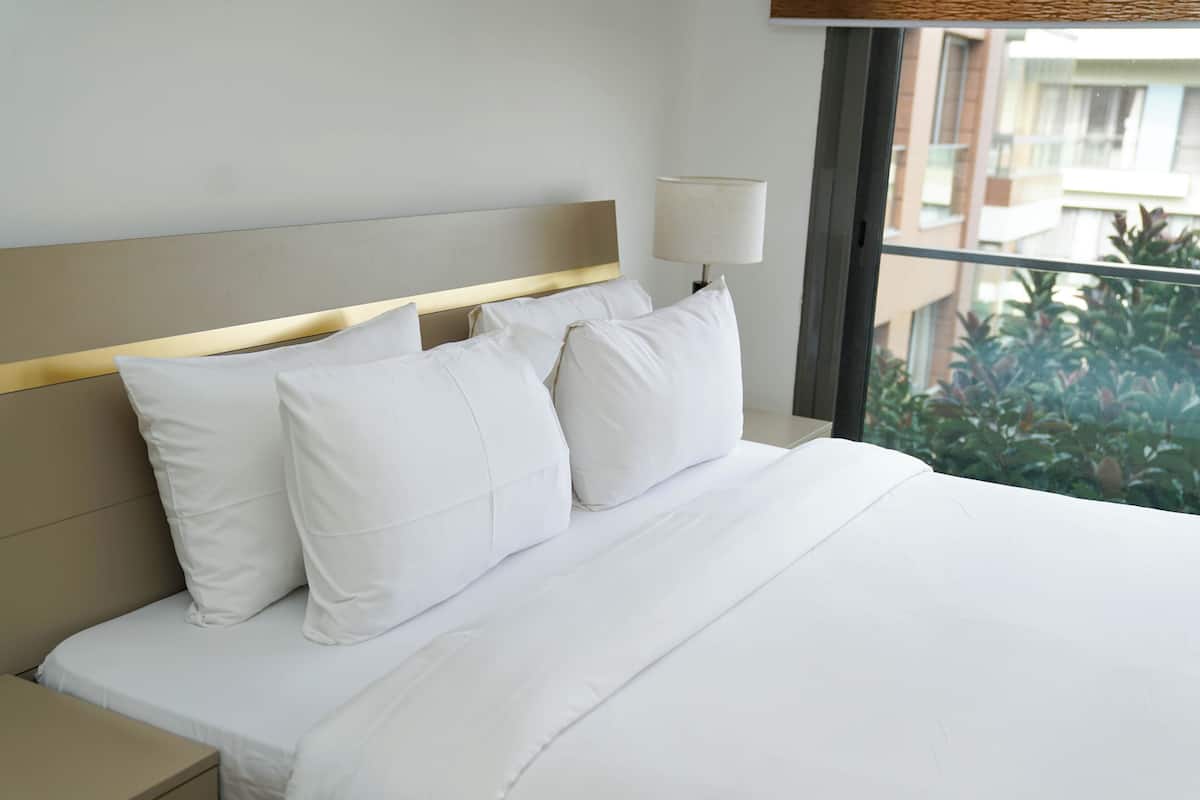 Een net opgemaakt bed met witte lakens en kussens, een nachtkastje en een lamp.