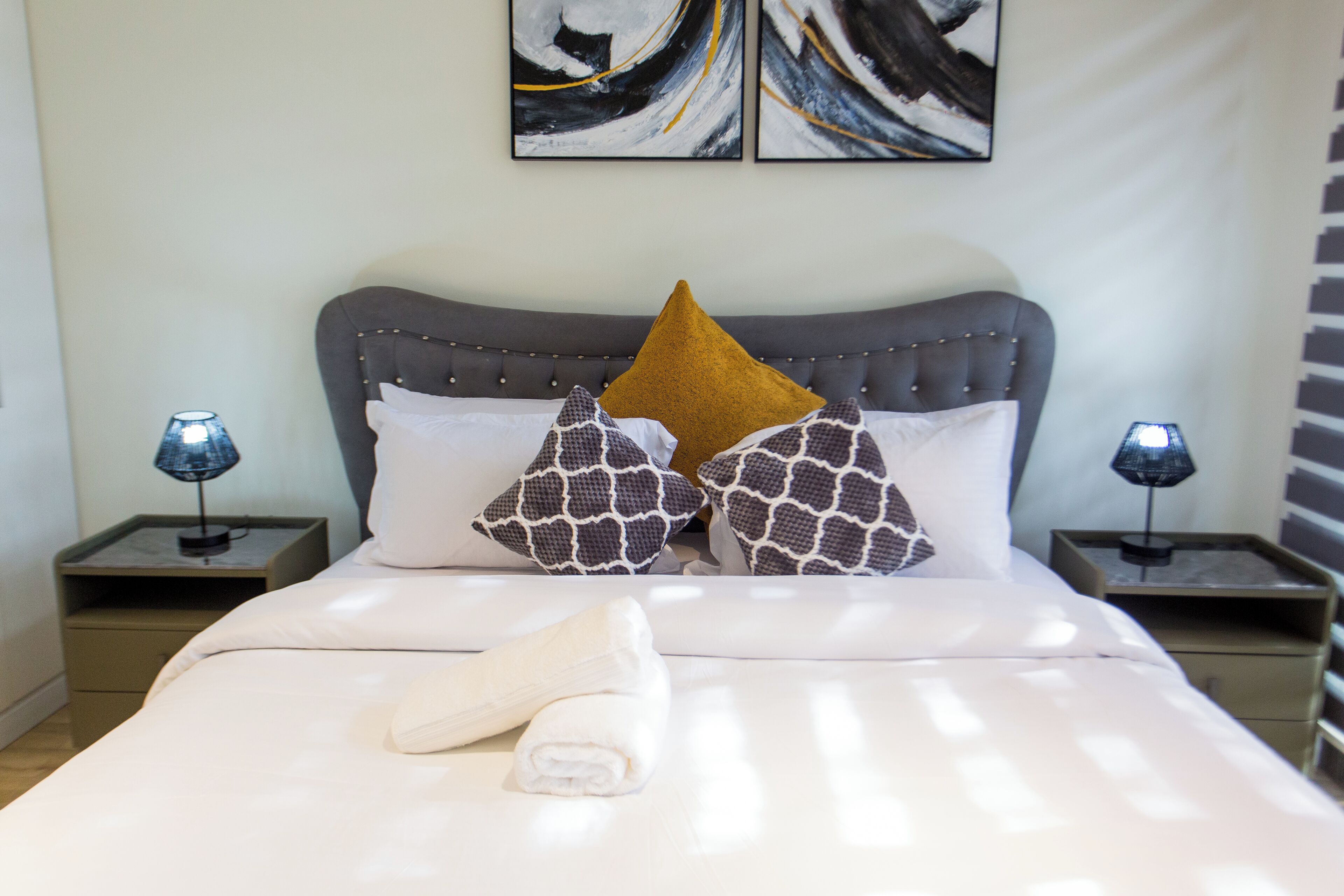 A neatly made bed with a grey upholstered headboard, two bedside lamps, and a pair of abstract wall art pieces.