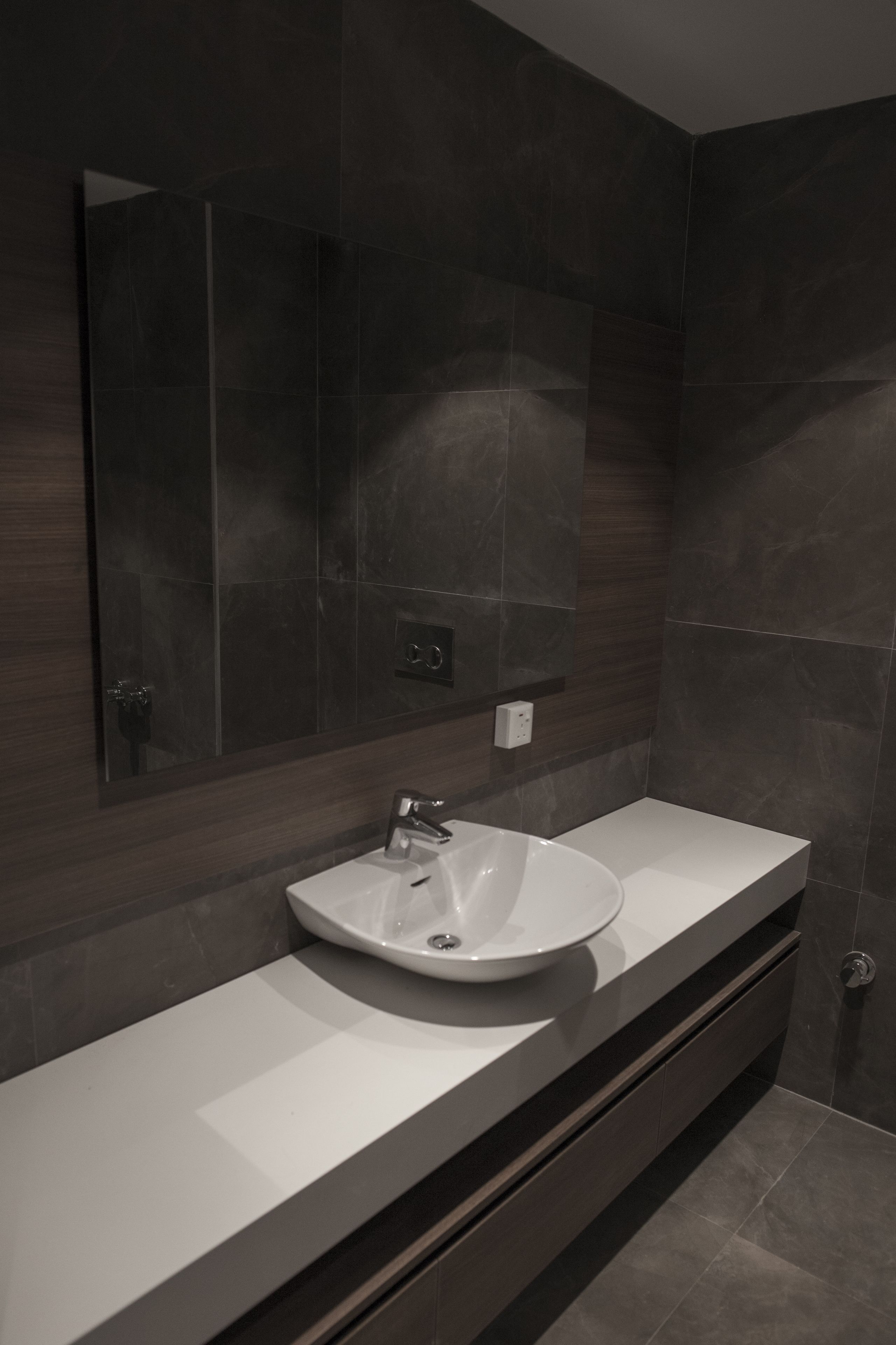 A modern bathroom with a large mirror, a white sink, and dark tiled walls.