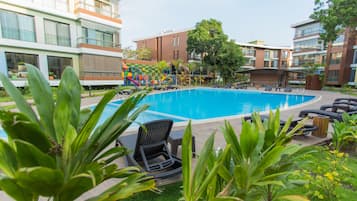 En swimmingpool omgivet af liggestole og grønt, med moderne bygninger i baggrunden.