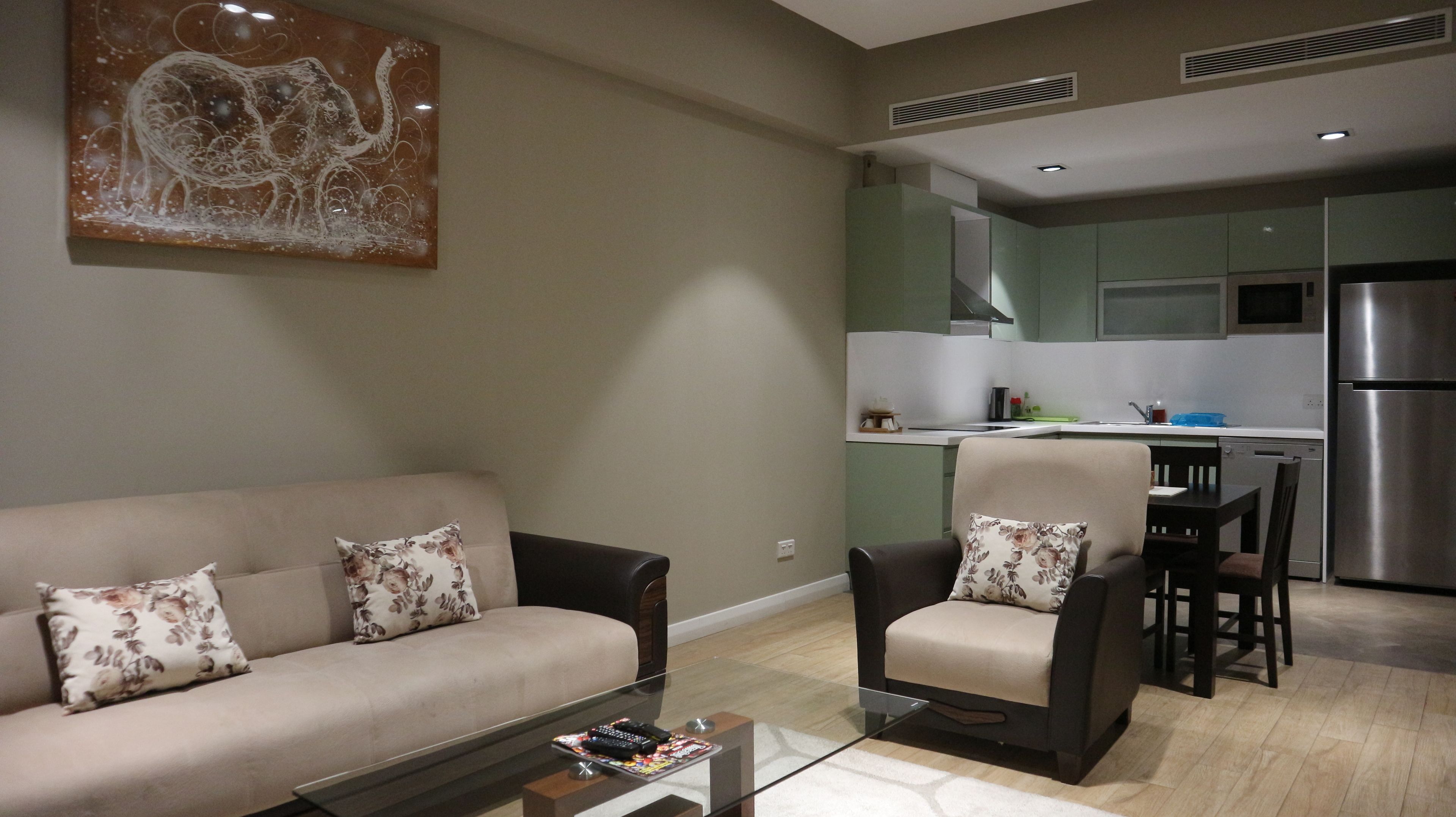 A living room with a sofa, armchairs, and a coffee table. A kitchen is visible in the background with a refrigerator and microwave.