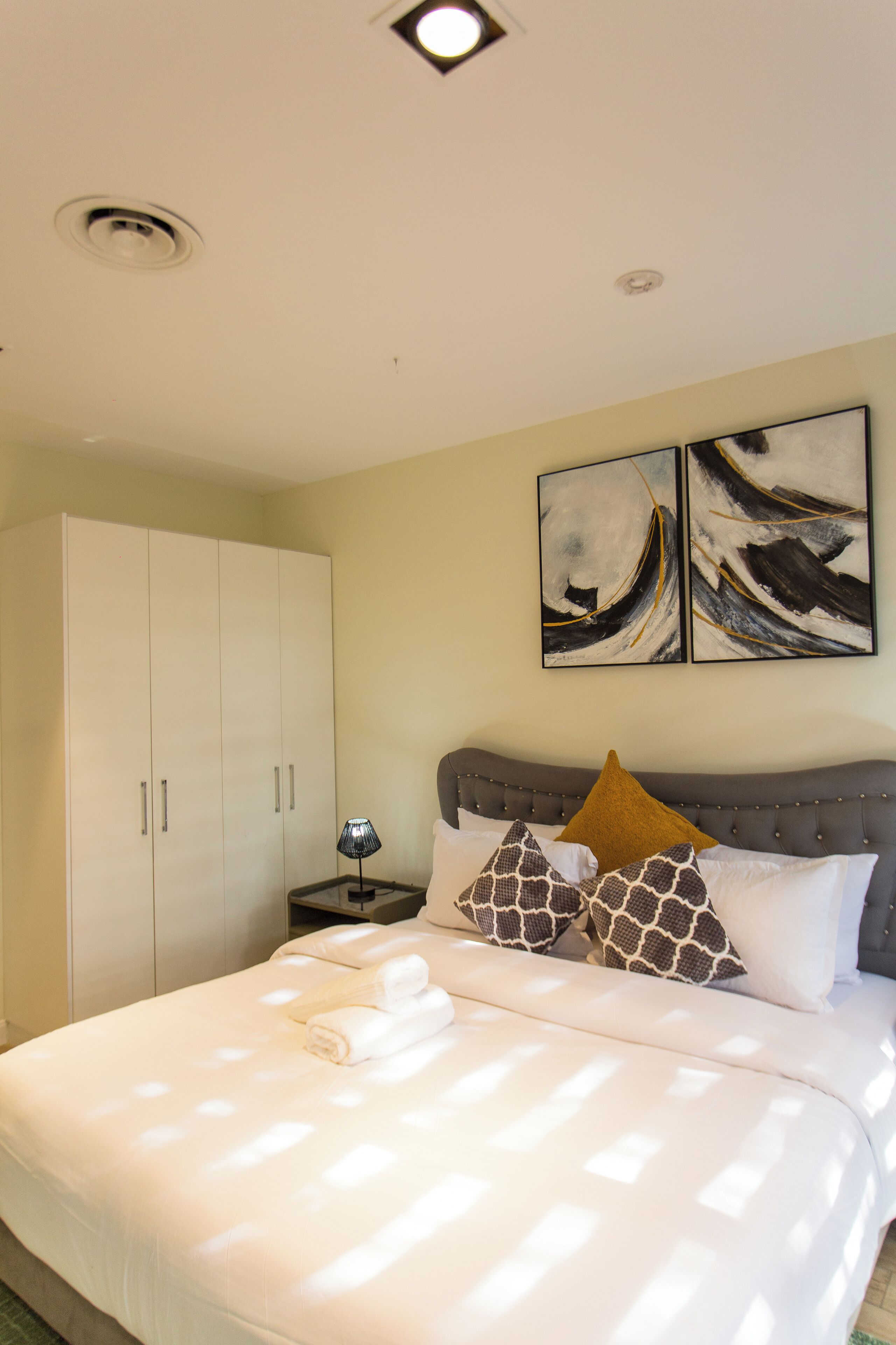 A neatly made bed with white bedding, a grey headboard, and decorative pillows. There is a white wardrobe to the left and two abstract paintings on the wall above the bed.
