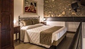 A bedroom with a stone wall, a bed with a quilted cover, a nightstand, a lamp, and a painting on the wall. - Isola Verde Resort (Castiglione del Lago)
