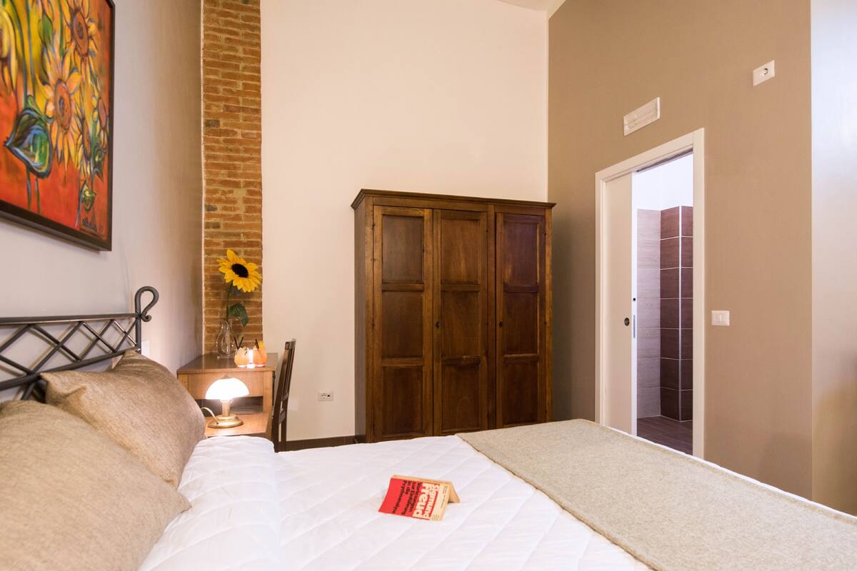 A bedroom with a bed, a wooden wardrobe, a nightstand with a lamp, and a painting on the wall.