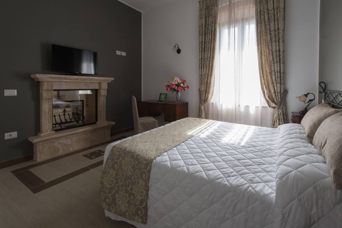 A hotel room with a large bed, a fireplace, a television, and a desk with a vase of flowers.