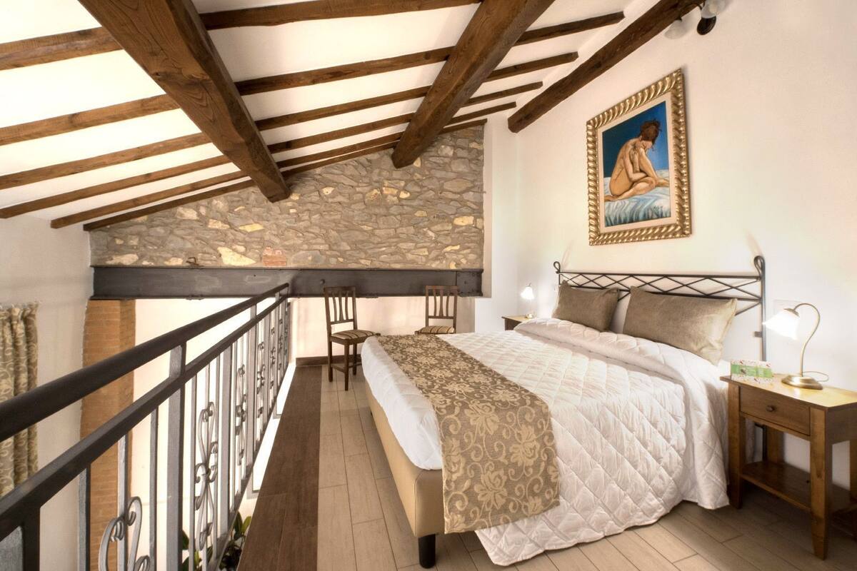 A bedroom with a stone wall, a bed with a patterned bedspread, a nightstand, a lamp, and a painting on the wall.