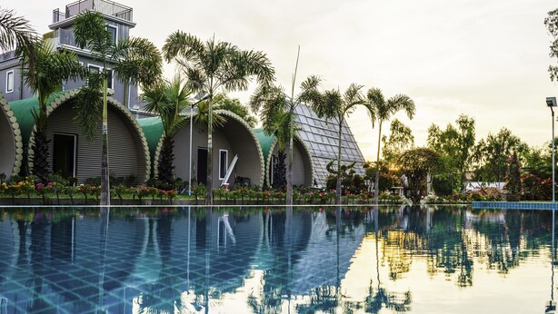 A swimming pool with a clear reflection of palm trees and a building with curved green roofs. - Garden Flower Resort (Siem Reap)