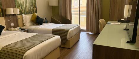 Habitación de hotel con dos camas, un escritorio, una silla y un amplio ventanal con cortinas.