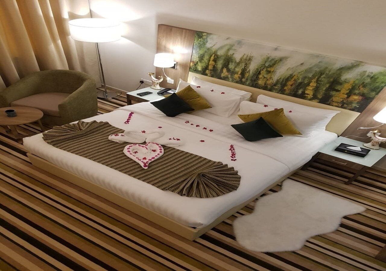 A hotel room with a large bed, a side table, a lamp, a chair, and a painting on the wall.