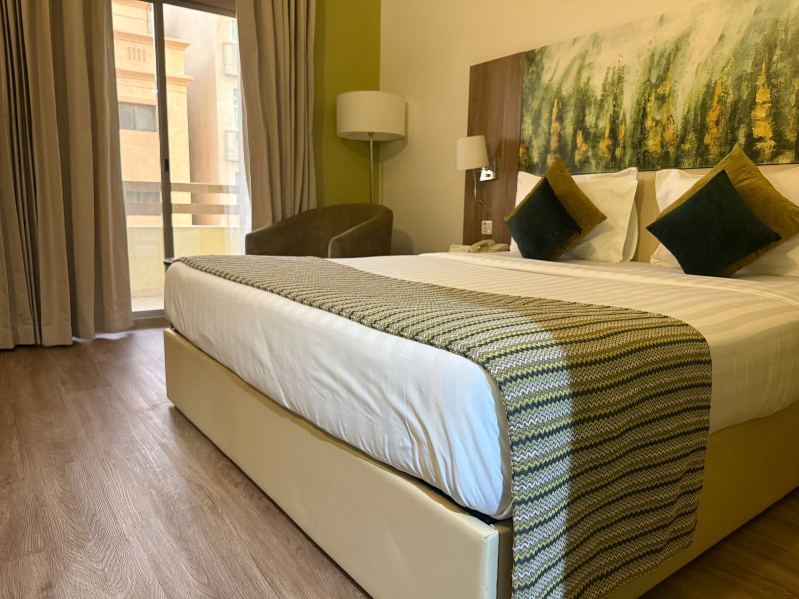 Ein Hotelzimmer mit einem großen Bett, einem Sessel, einem Bild an der Wand und Blick auf Gebäude.