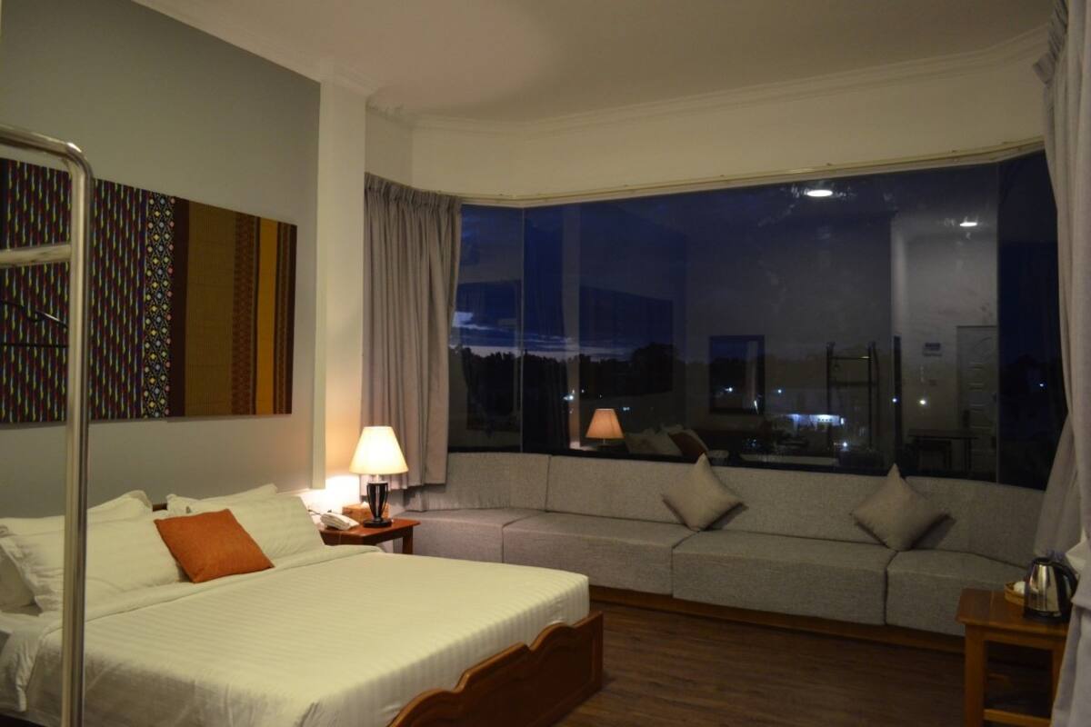 A hotel room with a large bed, a sofa, a nightstand, and a window with a city view.