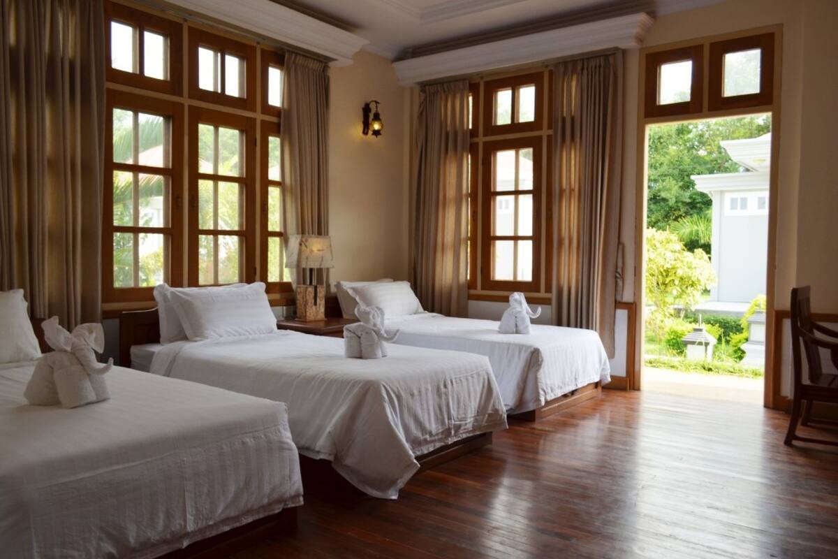 A hotel room with three beds, wooden flooring, large windows, and a view of greenery outside.