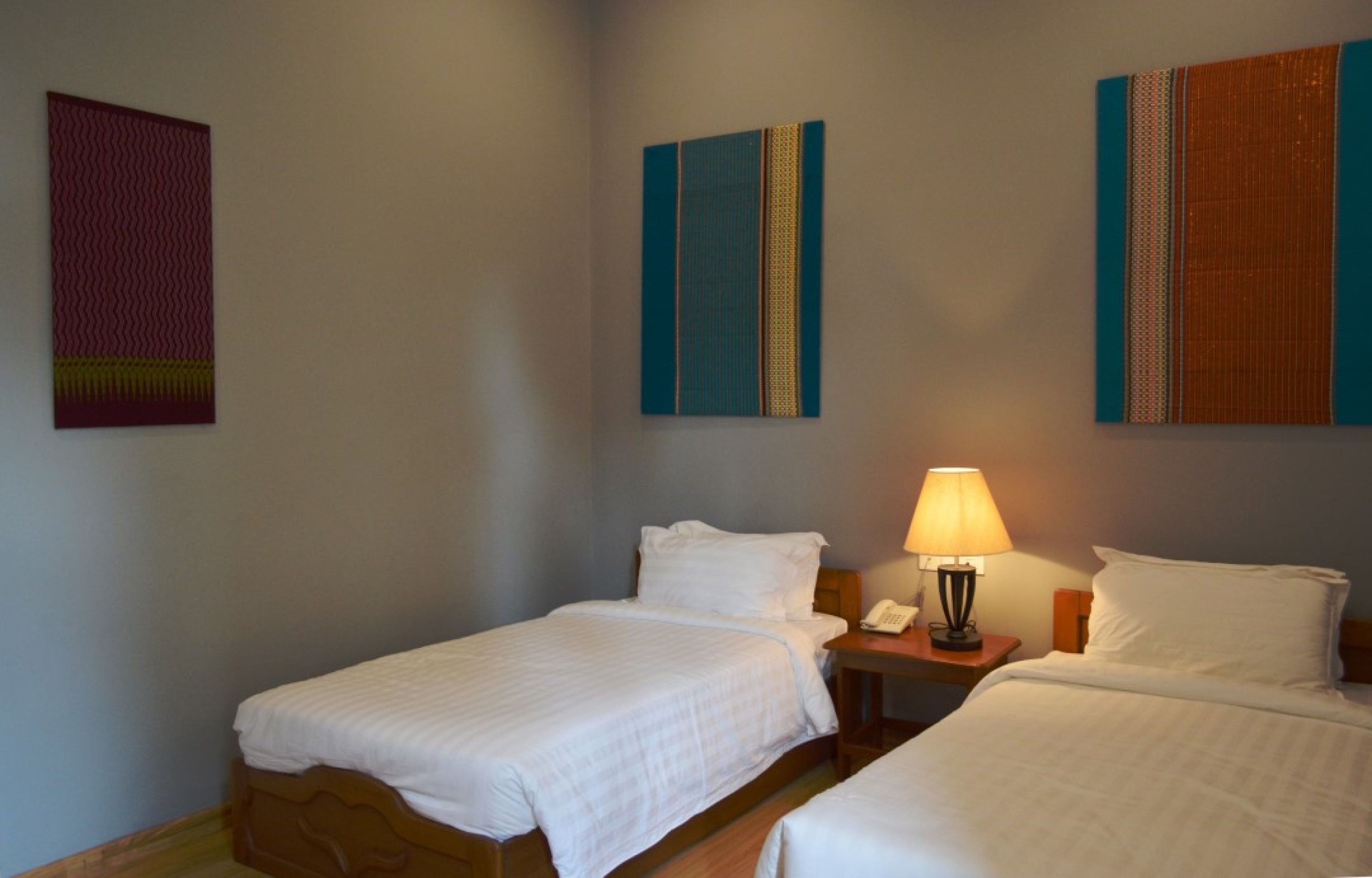 A hotel room with two single beds, a nightstand with a lamp, and artwork on the walls.