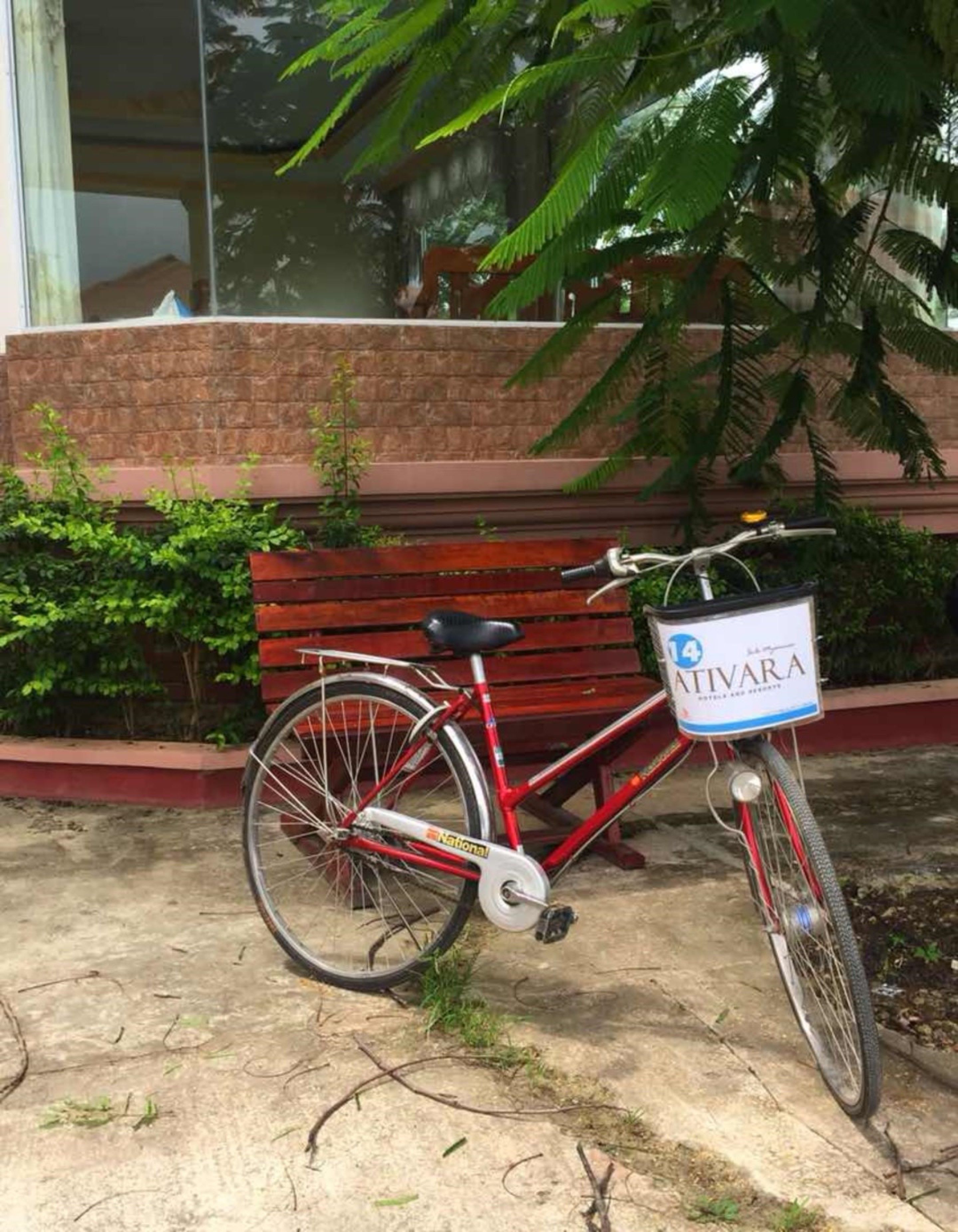 A red bicycle with a delivery box labeled "ATIVARA" parked in front of a building with a brick wall and a wooden bench.
