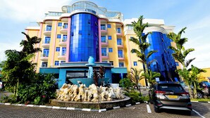 Un hotel a più piani con una facciata distintiva blu e gialla, una fontana centrale e un'area di parcheggio con auto.
