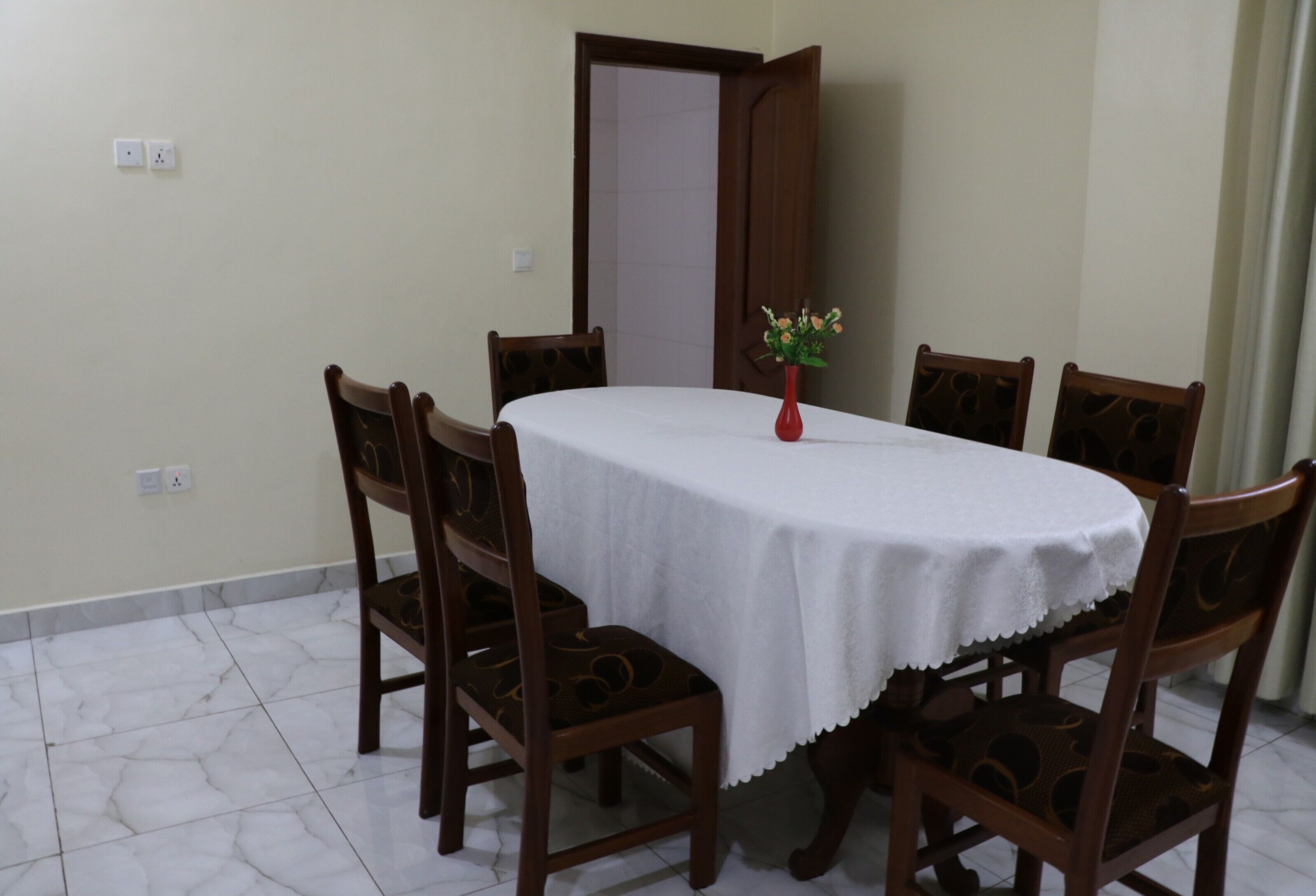 A dining area with a round table covered by a white tablecloth, surrounded by six wooden chairs with intricate back designs.
