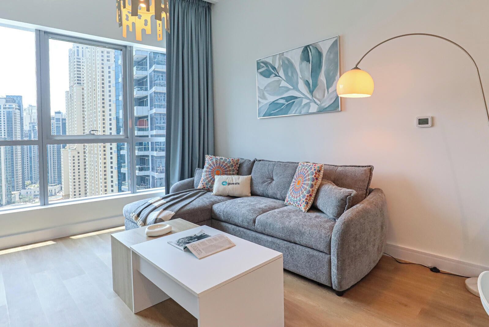 Un salon moderne avec un canapé gris, une table basse blanche, une vue sur la ville à travers de grandes fenêtres et un lampadaire.