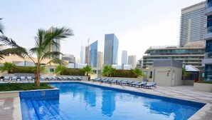 Une piscine sur le toit, avec des chaises longues, des palmiers et des bâtiments modernes en arrière-plan.