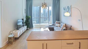 A modern living room with a sofa, TV, wooden table, and a view of tall buildings through large windows.