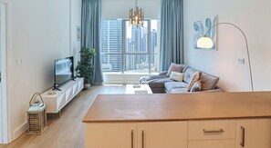 A modern living room with a sofa, TV, wooden table, and a view of tall buildings through large windows. - HiGuests - Bay central (Dubai)