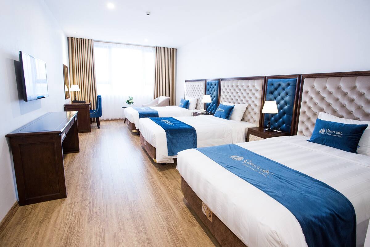 Une chambre d’hôtel avec trois lits, une télévision, un bureau et une grande fenêtre avec des rideaux.