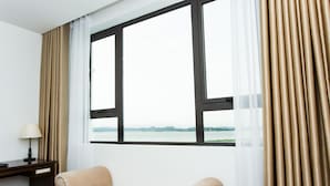 A hotel room with a large window, a sofa, a desk with a lamp, and a view of a body of water. - Song Loc Luxury (Bai Chay)