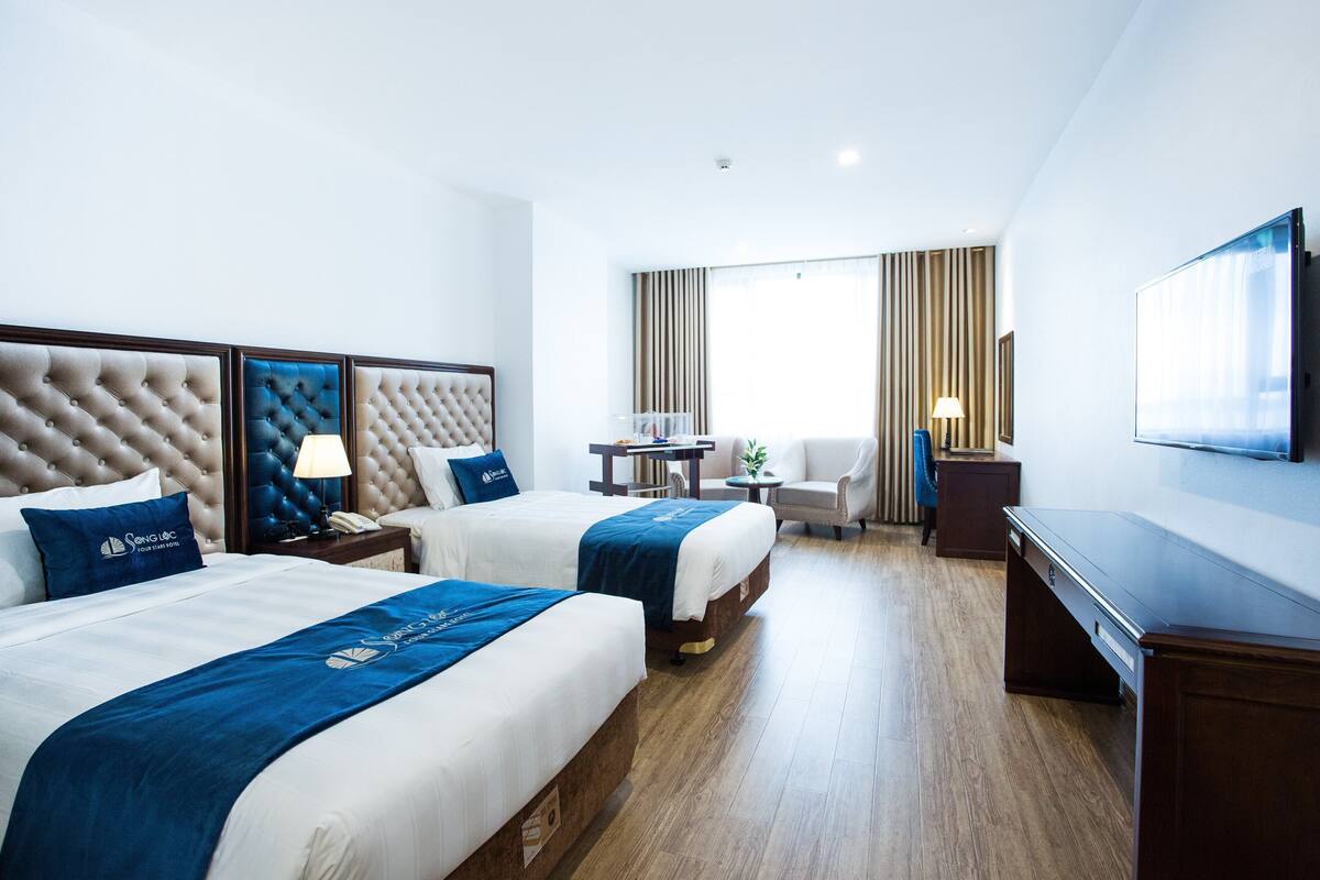Une chambre d’hôtel avec deux lits, un bureau, une chaise, une télévision et une fenêtre avec des rideaux.