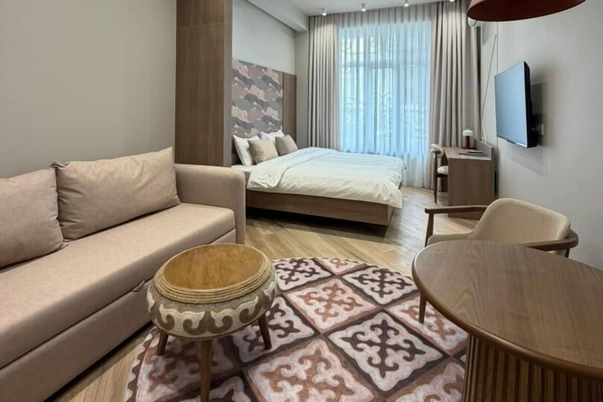 Una habitación de hotel moderna con sofá, una mesa redonda, alfombra estampada, cama, escritorio y televisor.