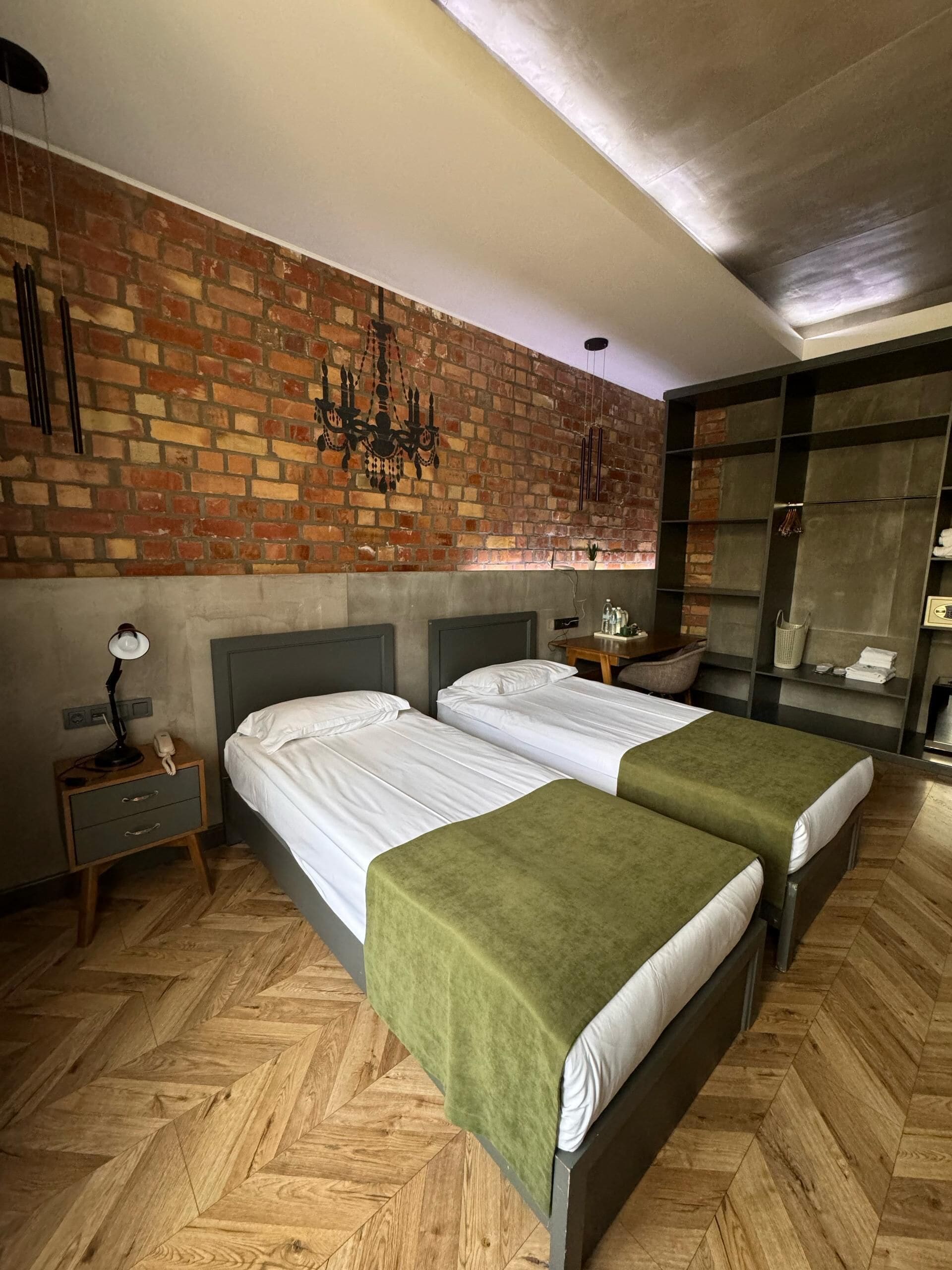 Une chambre d’hôtel avec deux lits, un mur en briques, un lustre et une armoire avec des étagères.