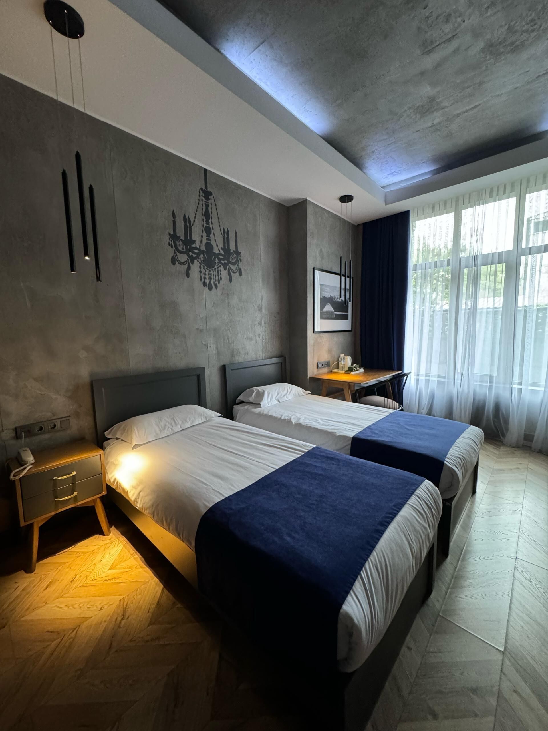 Una habitación de hotel moderna con dos camas, un escritorio, una araña y un amplio ventanal con cortinas.