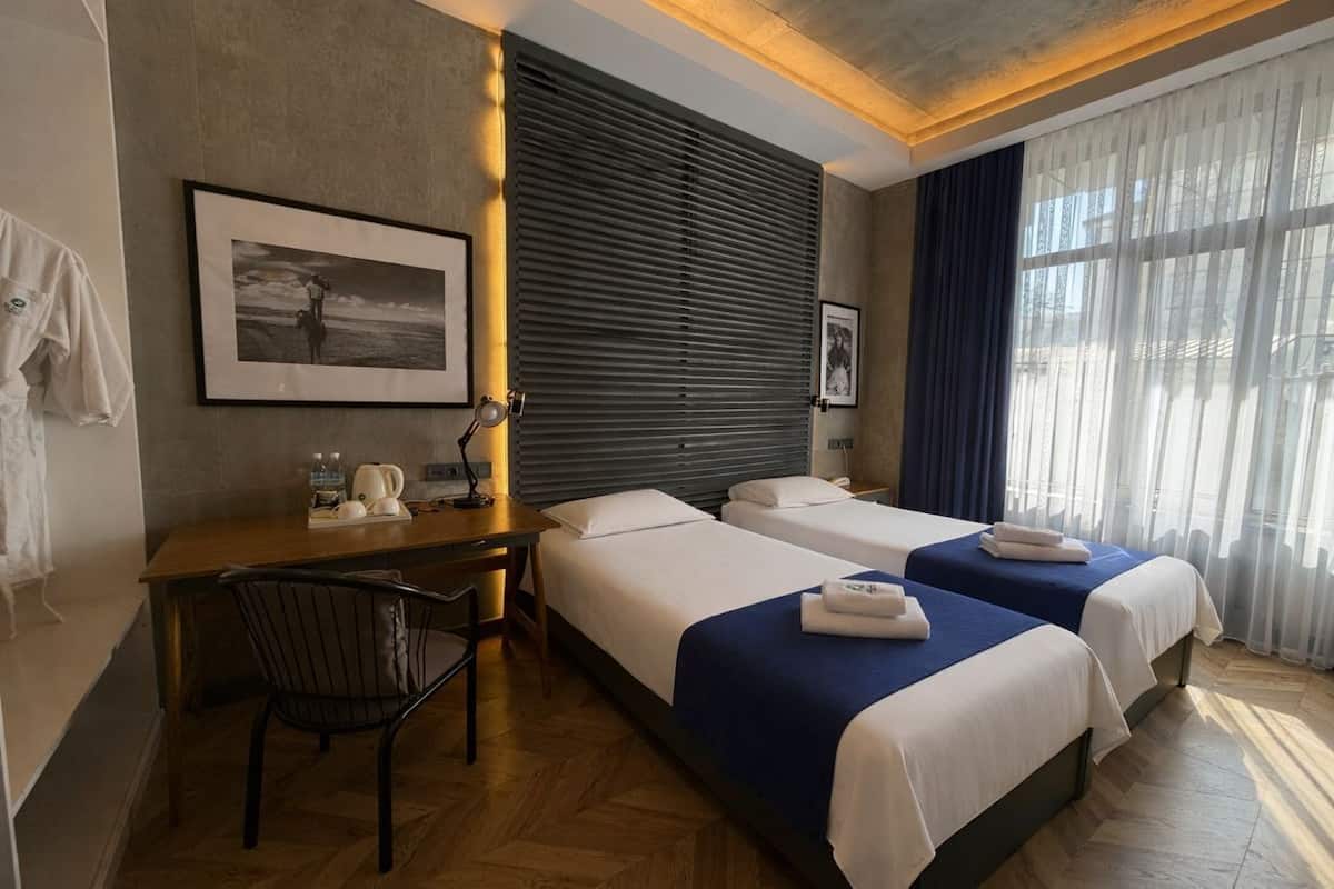 Habitación de hotel con dos camas, un escritorio, una silla y un ventanal con cortinas.