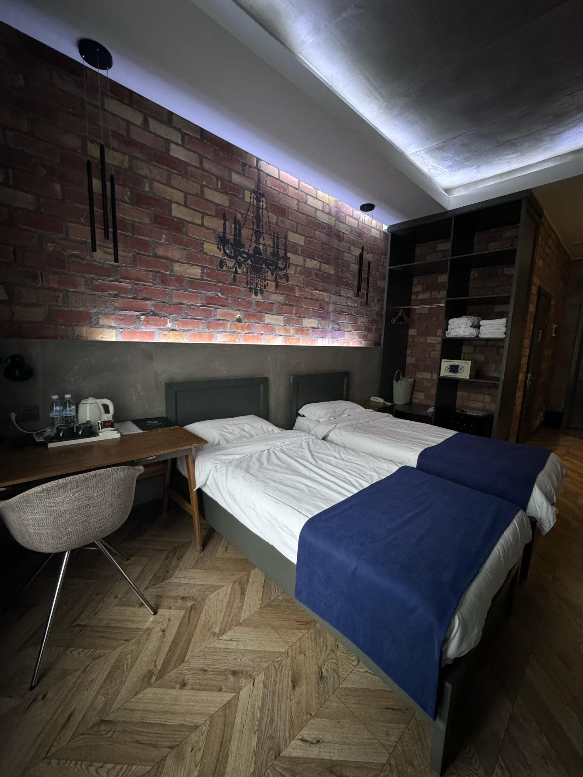 A bedroom with a brick wall, a bed with white and blue bedding, a desk with a chair, and a wardrobe.