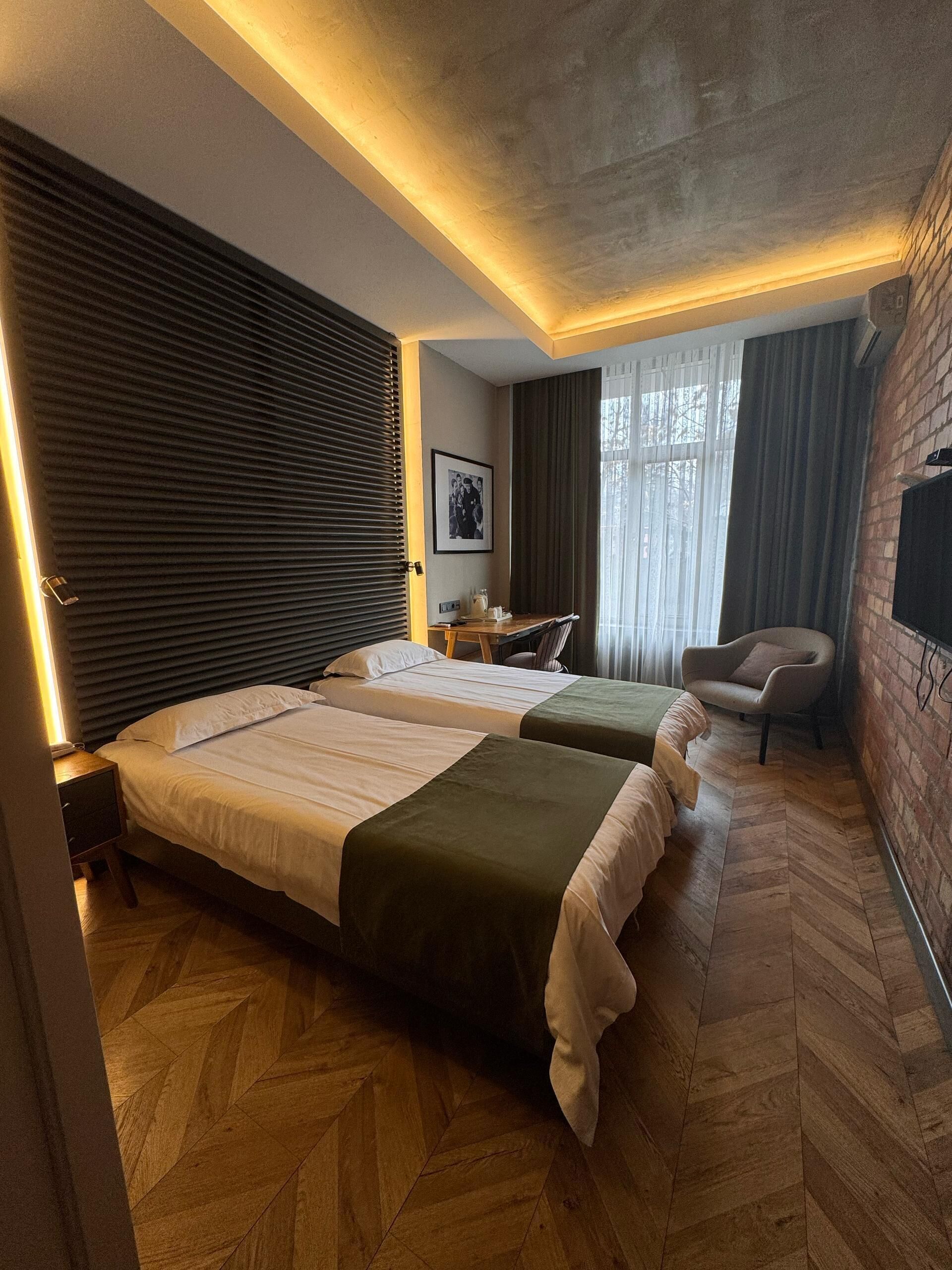 A hotel room with two beds, a desk, a chair, a picture on the wall, and a brick wall.