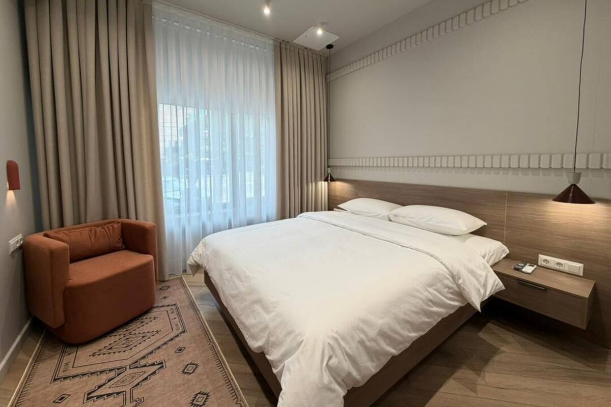 Habitación de hotel moderna con una cama grande, un sillón marrón, una mesita de noche con una lámpara y vistas a la ciudad desde la ventana.