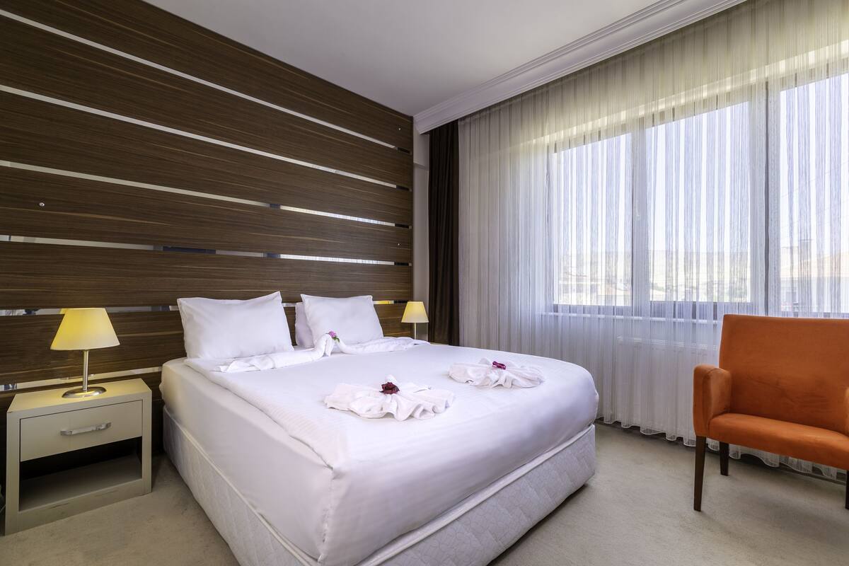 A hotel room with a large bed, two bedside tables with lamps, a wooden headboard, a large window with sheer curtains, and an orange armchair.