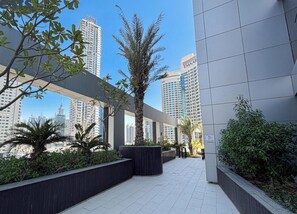 A modern urban walkway with tall buildings, planters with greenery, and a clear sky.