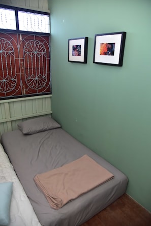 A single bed with a grey mattress and a beige blanket, a window with decorative metal bars, and two framed pictures on the wall.